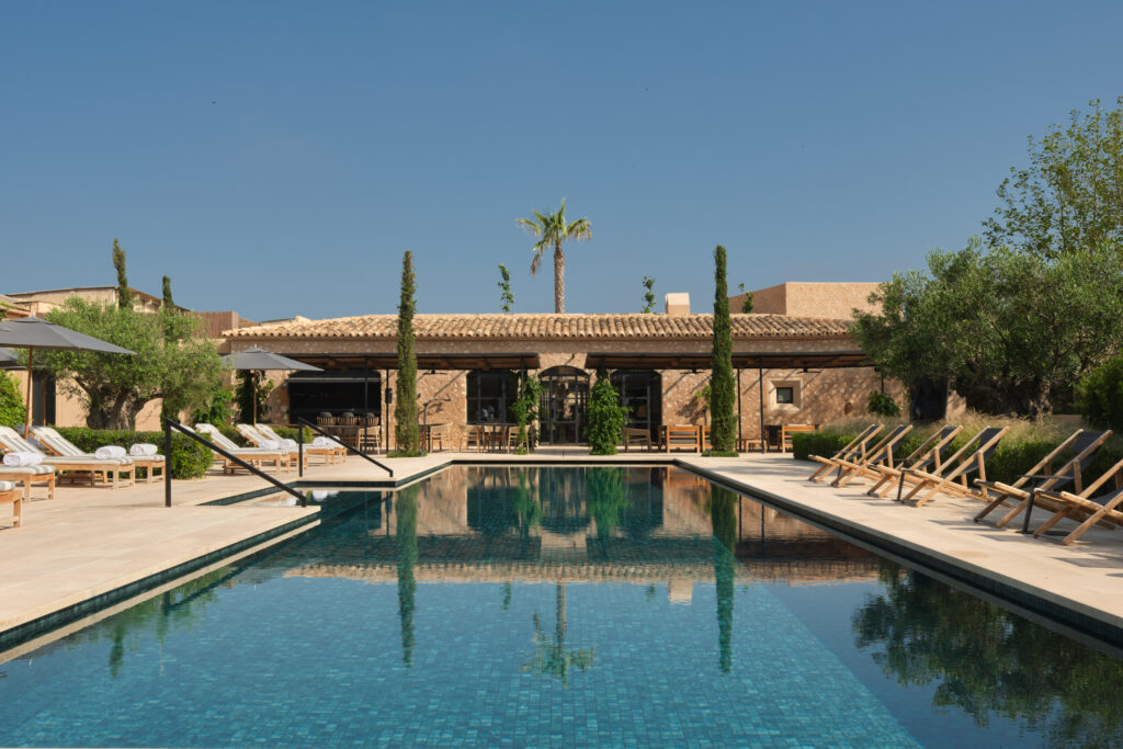A serene outdoor pool area at Can Ferrereta, featuring loungers, sunshades, and the beautifully restored 17th-century mansion in the background, surrounded by greenery and clear blue skies.